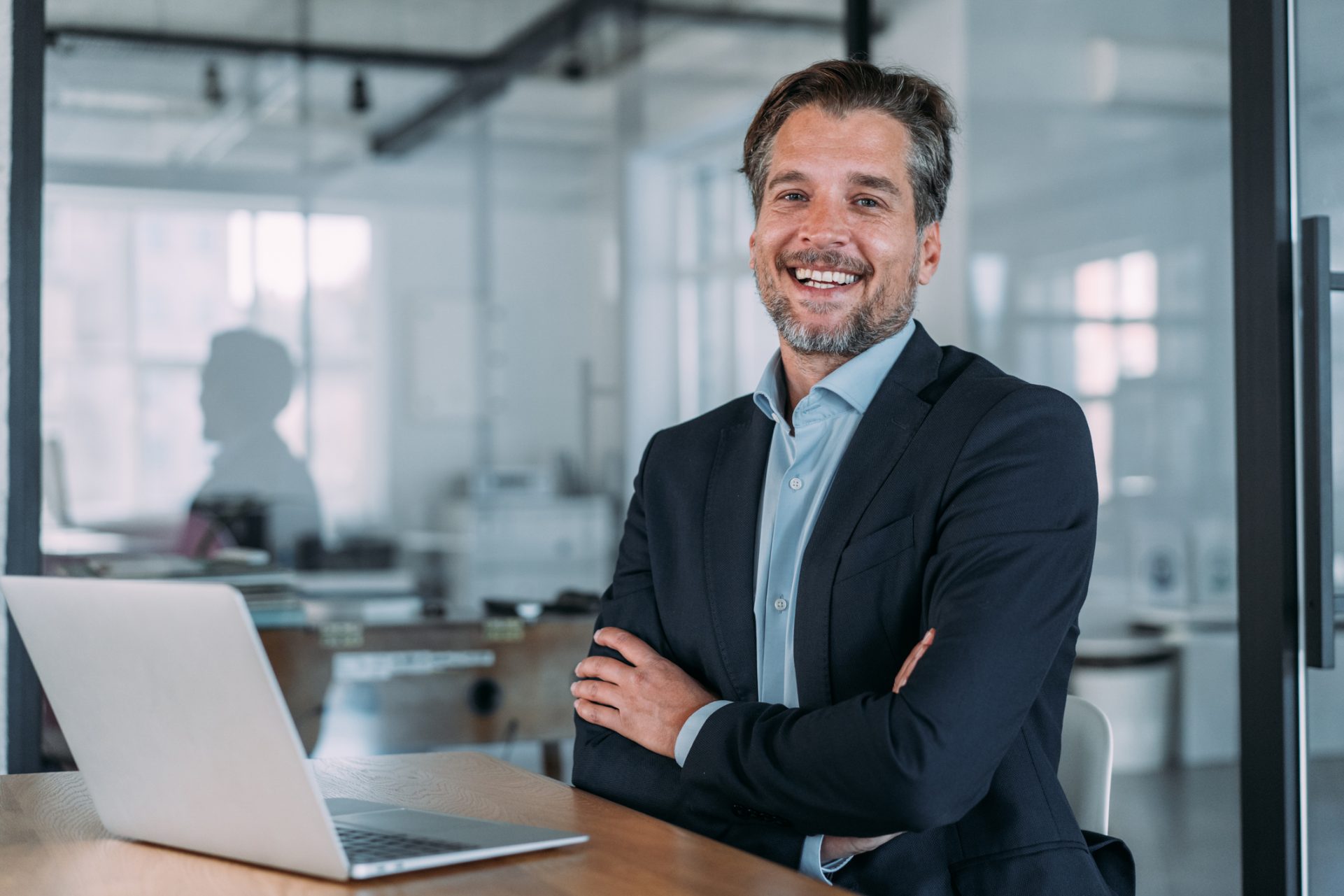 Shot of a businessman sitting on desk in modern office and working on laptop. Elegant handsome businessman sitting in his office and using laptop.